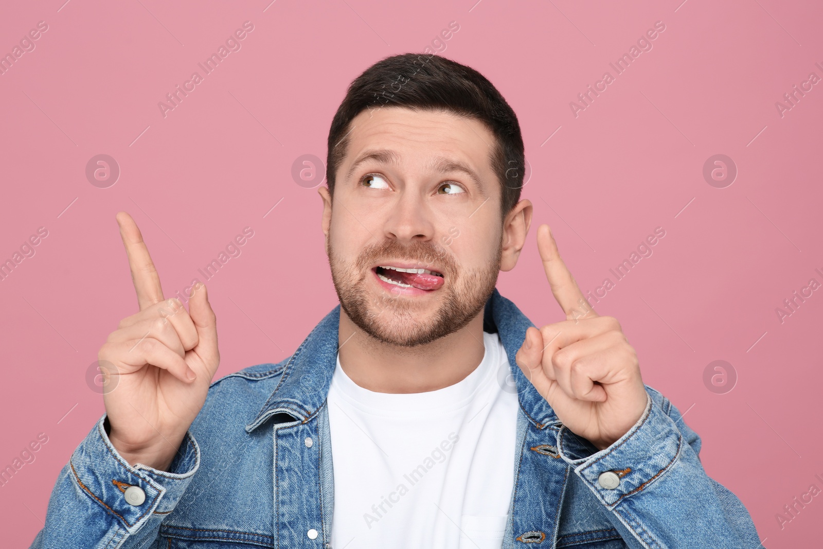 Photo of Happy man showing his tongue and pointing at something on pink background