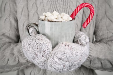Photo of Woman in knitted mittens holding cup of delicious hot chocolate with marshmallows and candy cane, closeup