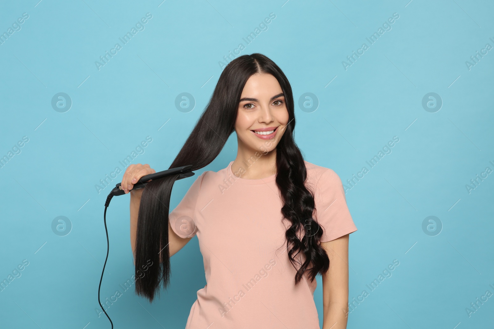 Photo of Beautiful happy woman using hair iron on light blue background