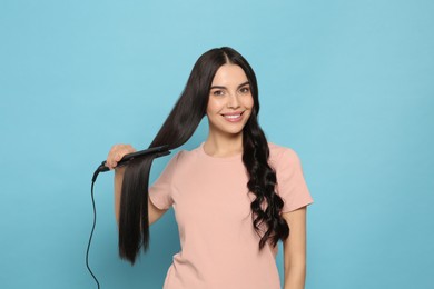 Beautiful happy woman using hair iron on light blue background