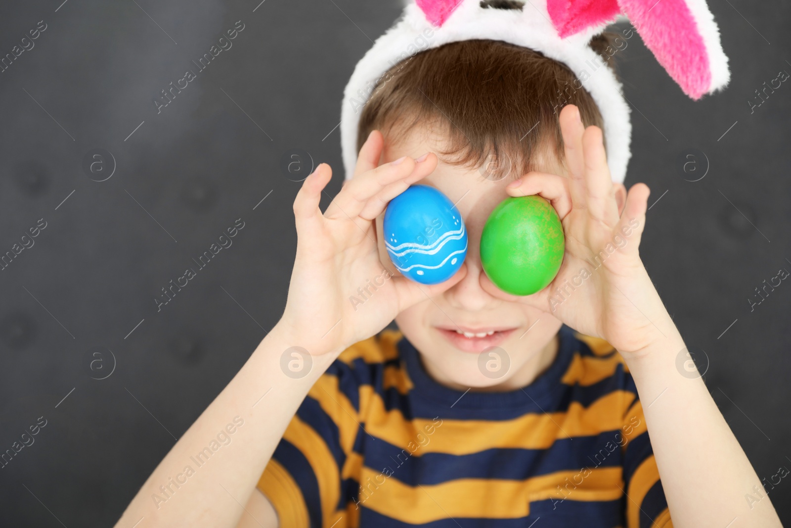 Photo of Little boy in bunny ears headband holding Easter eggs near eyes at home, closeup