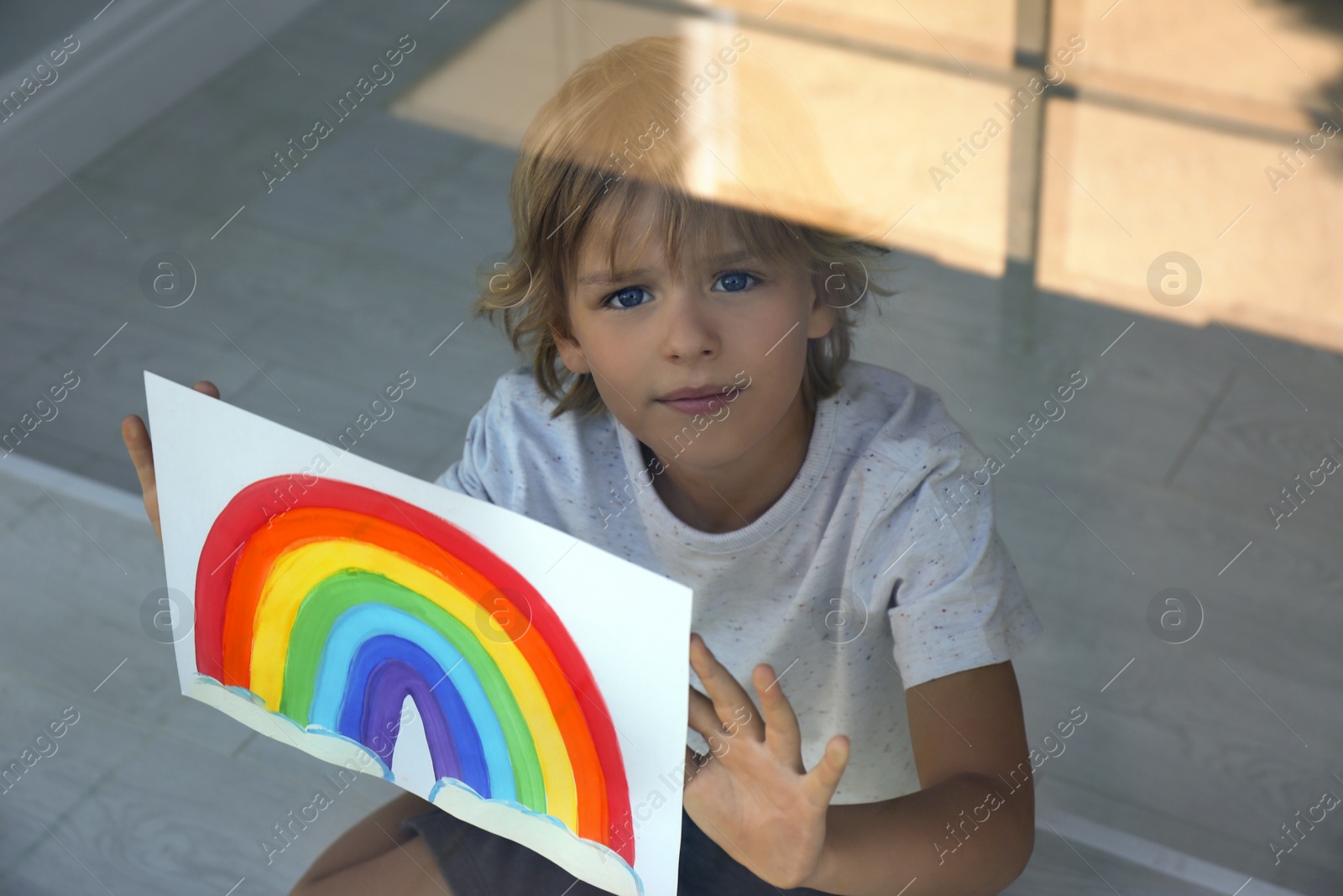 Photo of Little boy with picture of rainbow near window, view from outdoors. Stay at home concept