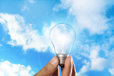 Image of Solar energy concept. Man holding glowing light bulb against blue sky with clouds, closeup