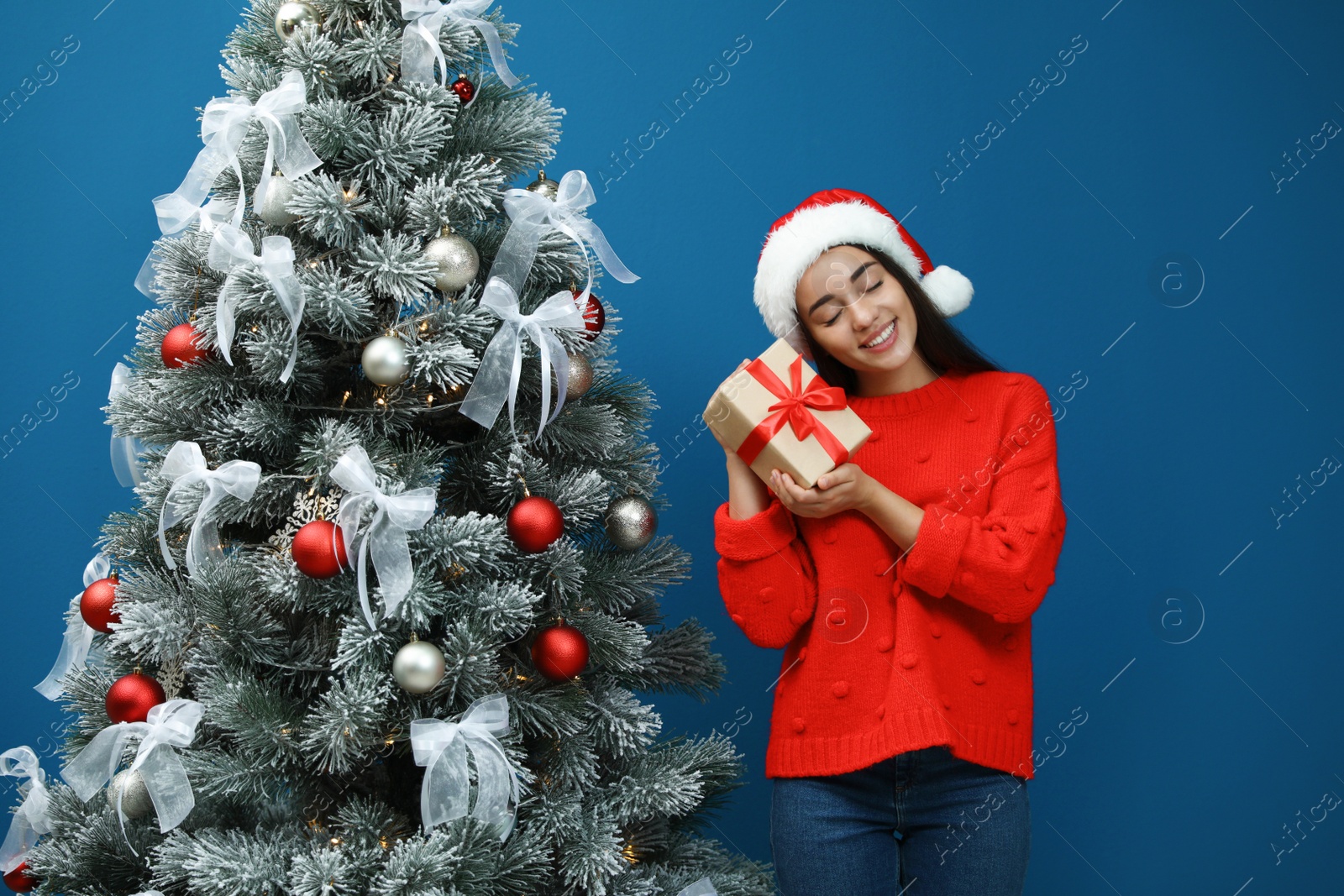 Image of Happy young woman in Santa hat with gift near Christmas tree on blue background