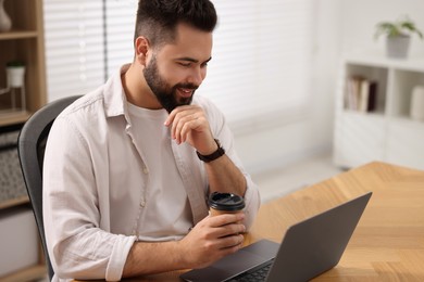 Photo of Young man with cup of coffee watching webinar at table in room