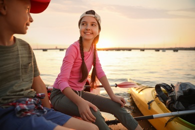 Happy children sitting on kayak near river at sunset. Summer camp
