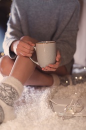 Woman with cup of hot beverage and book at home in winter evening, closeup