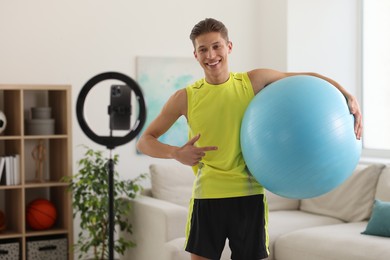 Photo of Smiling sports blogger holding fit ball while streaming online fitness lesson at home