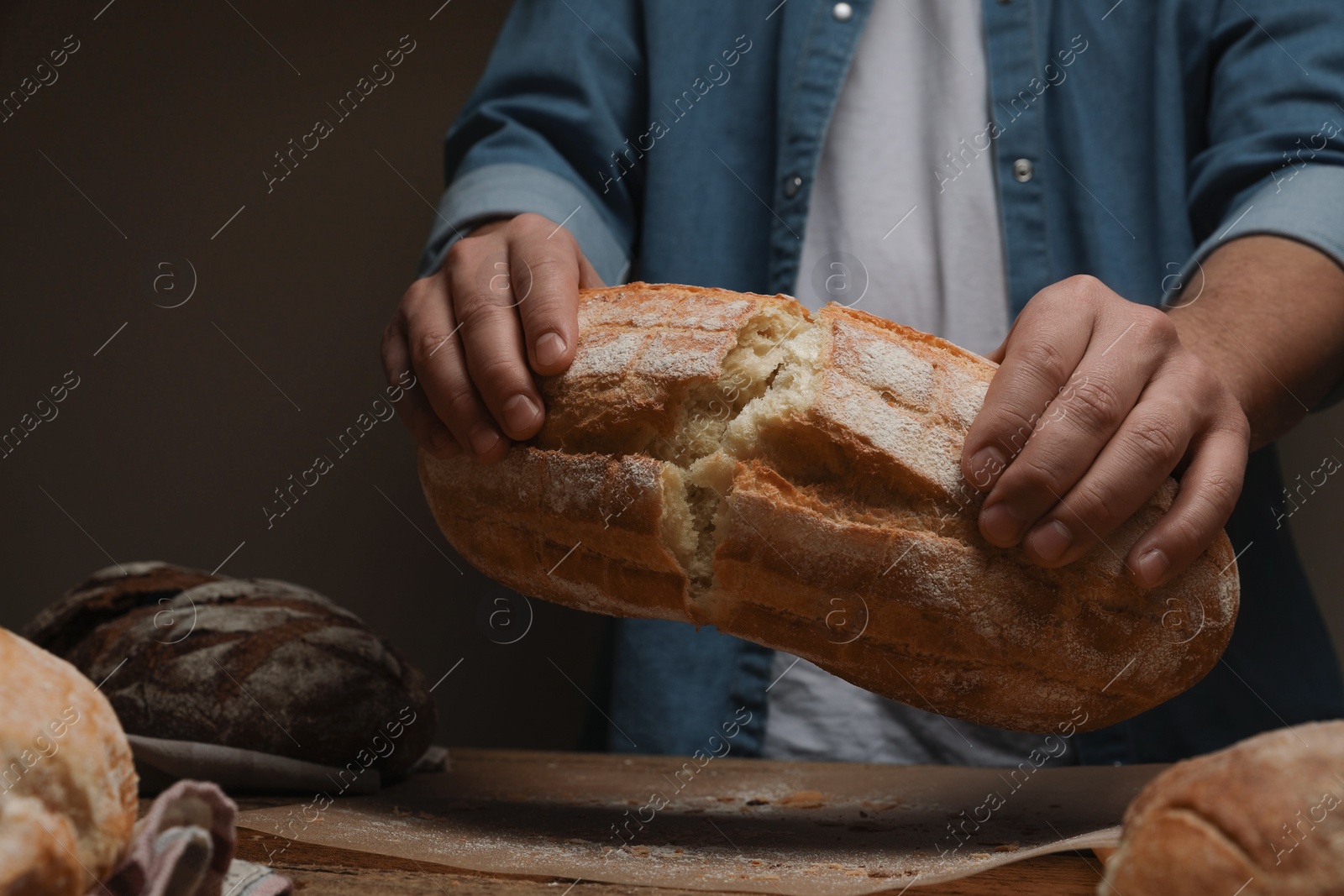 Photo of Man breaking loaf of fresh bread at wooden table, closeup