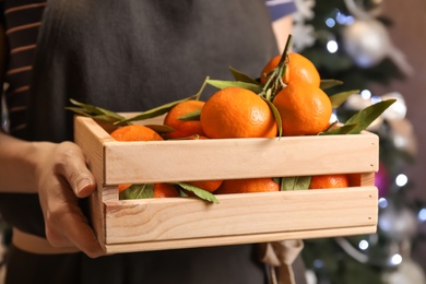 Photo of Woman with wooden crate of ripe tangerines and Christmas tree on background, closeup
