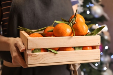 Woman with wooden crate of ripe tangerines and Christmas tree on background, closeup