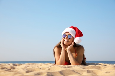 Beautiful young woman in Santa hat and bikini on beach, space for text. Christmas vacation