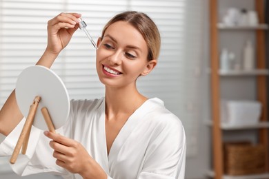 Beautiful woman applying cosmetic serum onto her face in front of mirror in bathroom, space for text