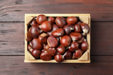 Fresh sweet edible chestnuts in crate on wooden table, top view