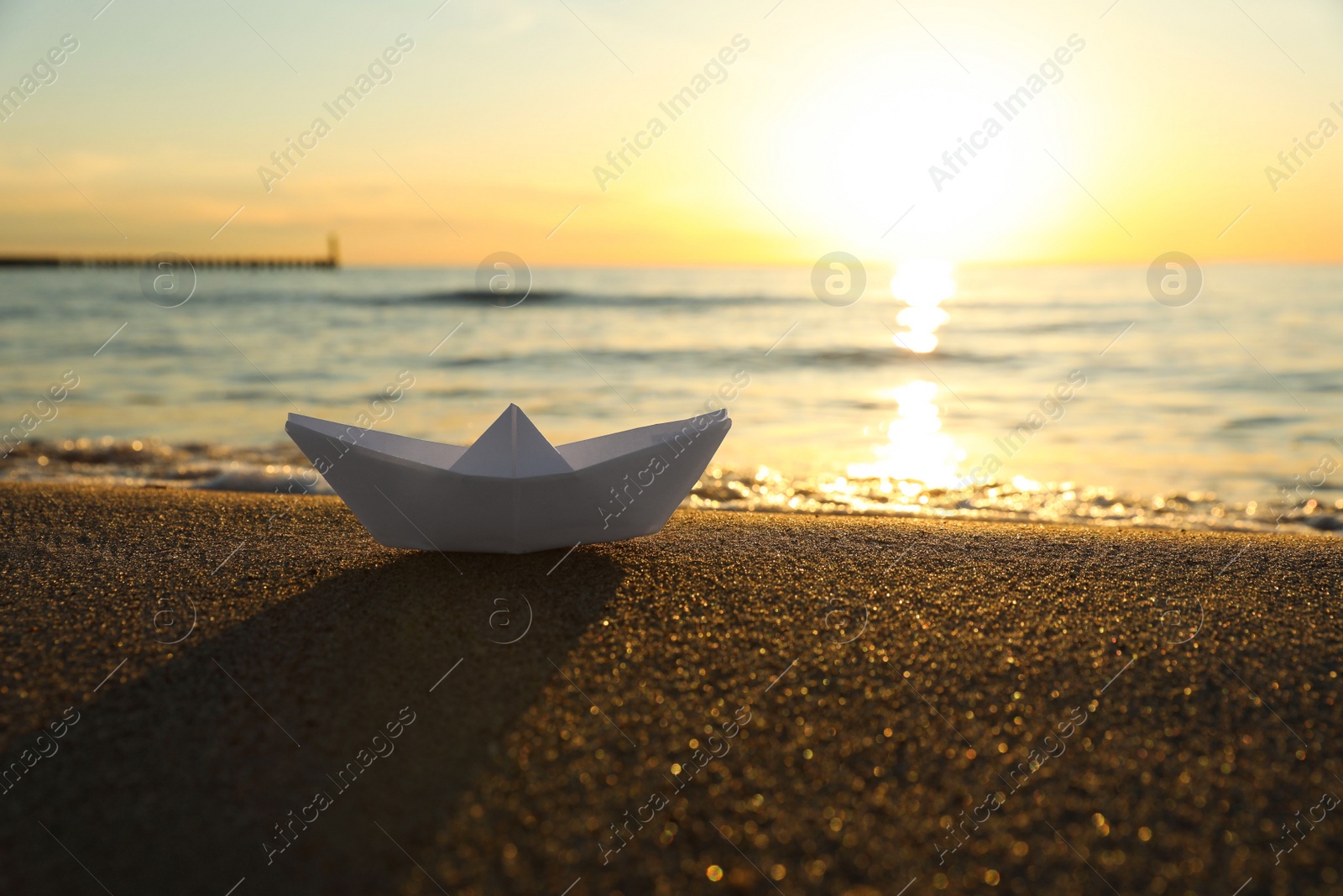 Photo of White paper boat on sand near sea at sunset. Space for text