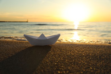 Photo of White paper boat on sand near sea at sunset. Space for text