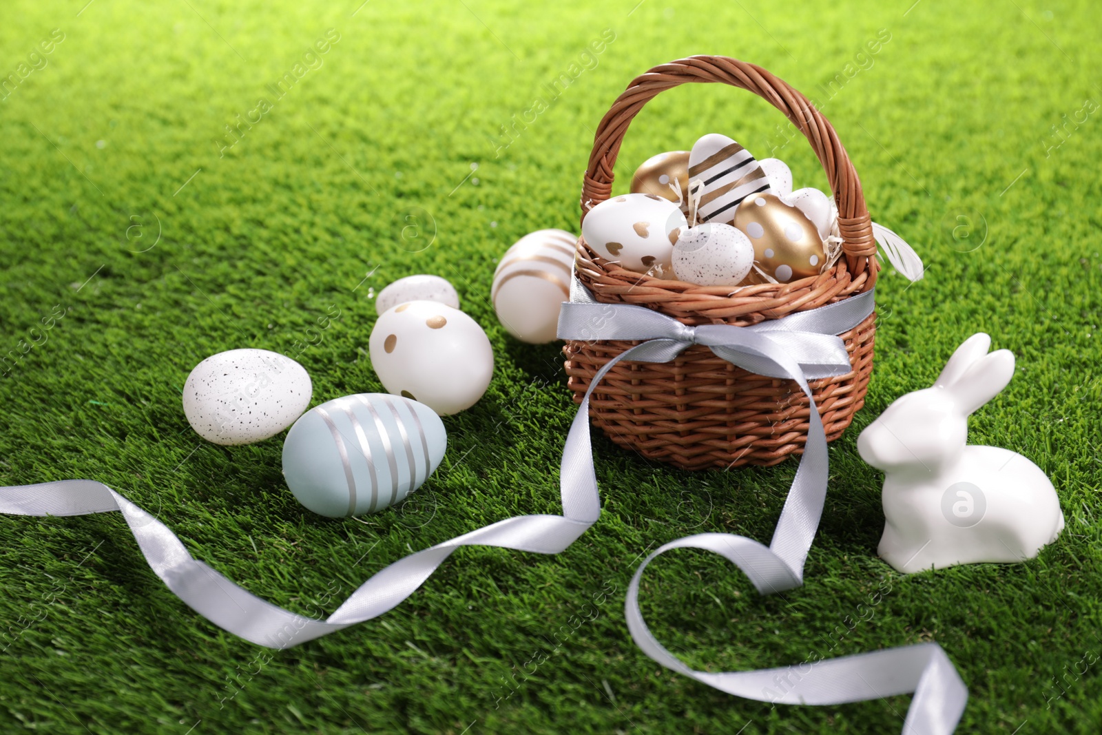Photo of Easter basket with beautifully painted eggs, ribbon and figure of rabbit on green grass