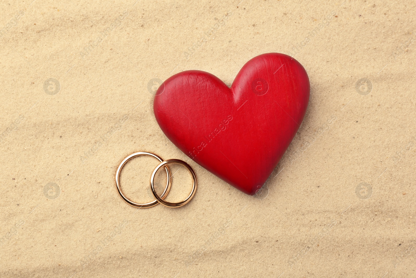 Photo of Honeymoon concept. Two golden rings and red wooden heart on sand, top view