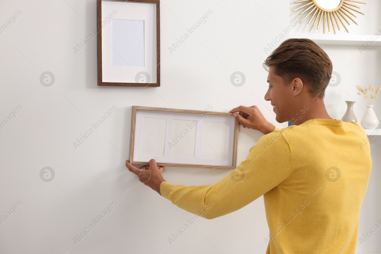 Photo of Young man hanging picture frames on white wall indoors