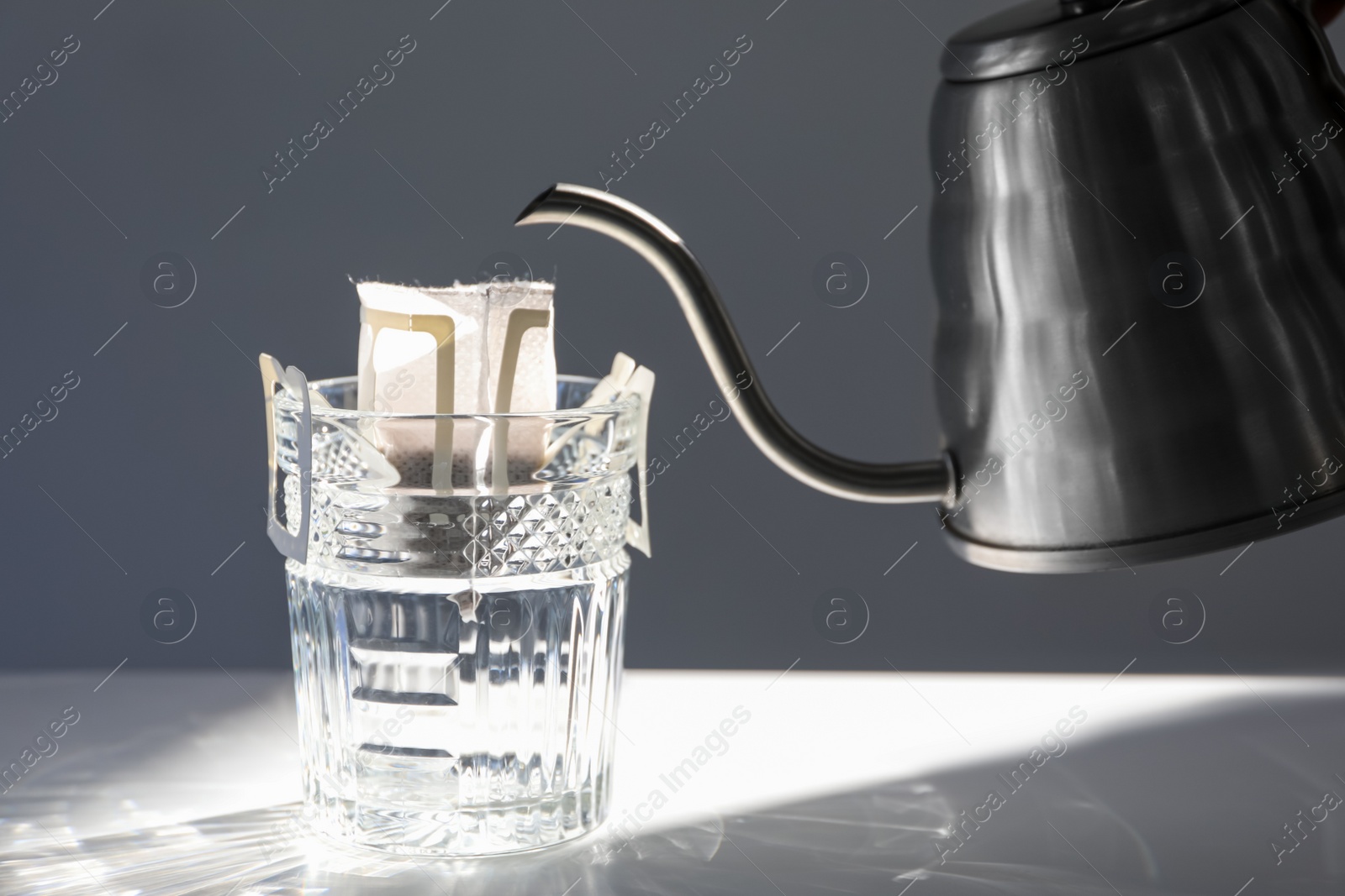 Photo of Glass with drip coffee bag and metal kettle on light grey table, closeup