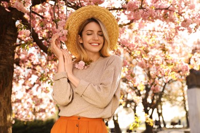 Young woman wearing stylish outfit near blossoming sakura in park. Fashionable spring look