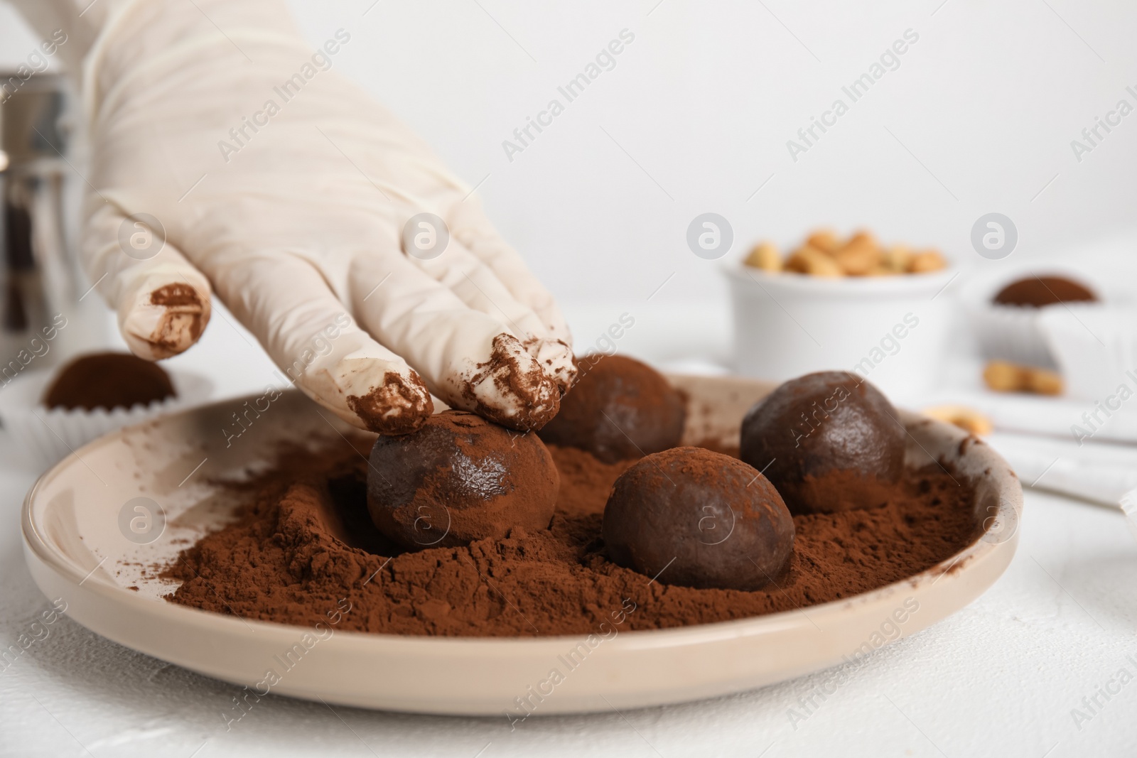 Photo of Confectioner covering delicious chocolate truffles with cocoa powder at white table, closeup