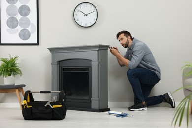 Photo of Man with screwdriver installing electric fireplace near wall in room