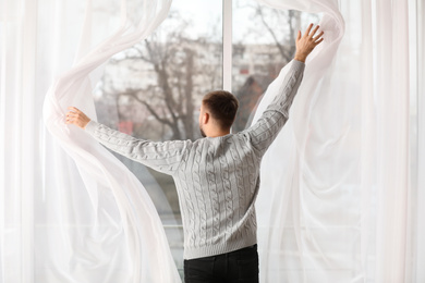 Photo of Young man opening window curtains at home, back view