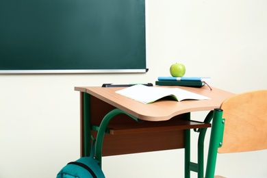 Photo of Wooden school desk with stationery, apple and backpack near chalkboard in classroom