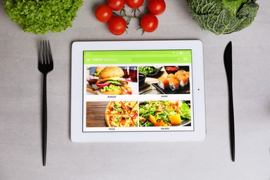 Photo of Modern tablet with open page for online food ordering, vegetables and cutlery on grey table, flat lay. Concept of delivery service
