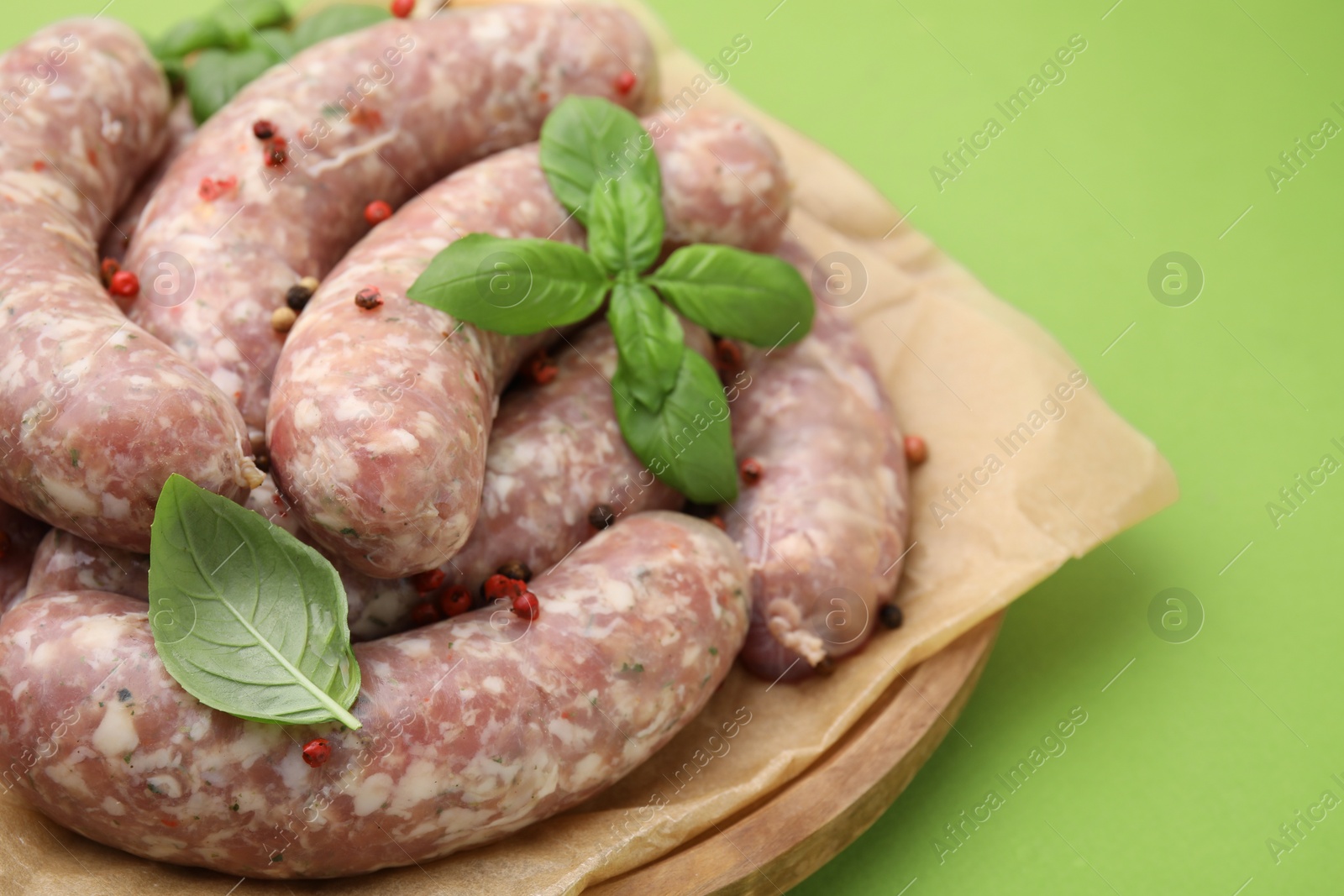 Photo of Board with raw homemade sausages, basil leaves and peppercorns on green table, closeup. Space for text