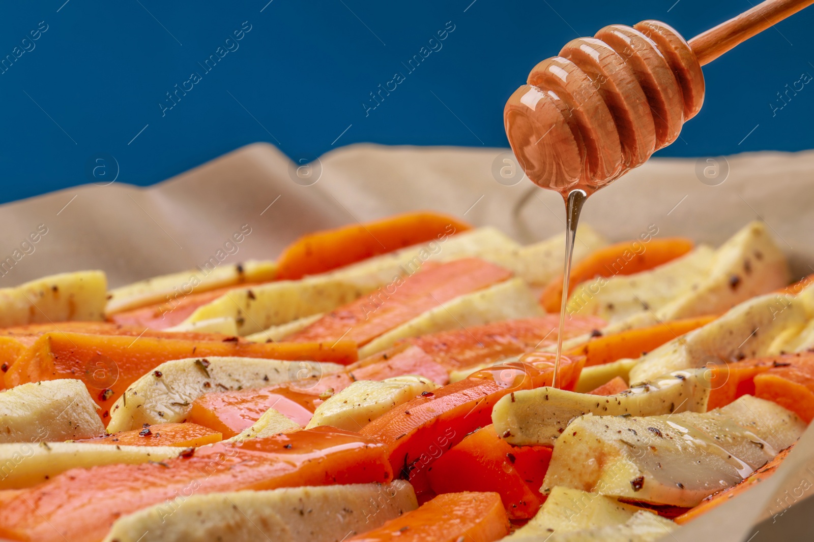 Photo of Pouring honey onto slices of parsnip and carrot against blue background, closeup