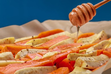 Photo of Pouring honey onto slices of parsnip and carrot against blue background, closeup