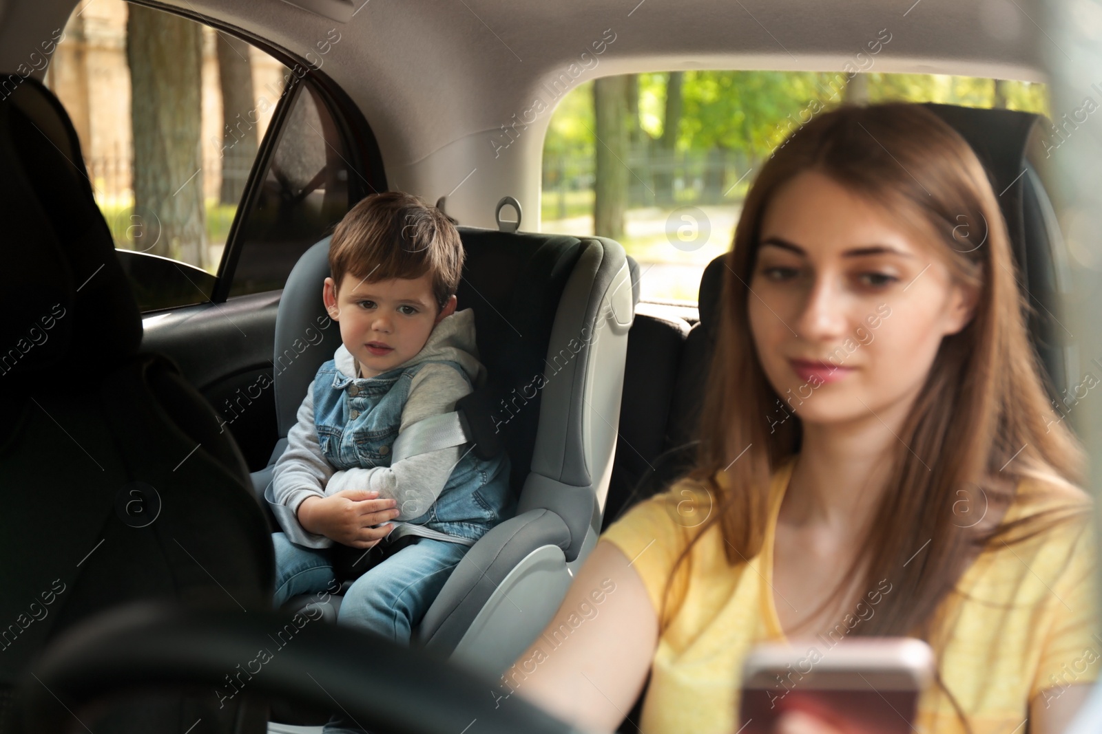 Photo of Mother using phone while driving car with her son on backseat. Child in danger