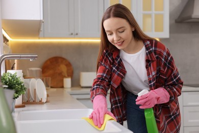 Photo of Woman with spray bottle and microfiber cloth cleaning sink in kitchen