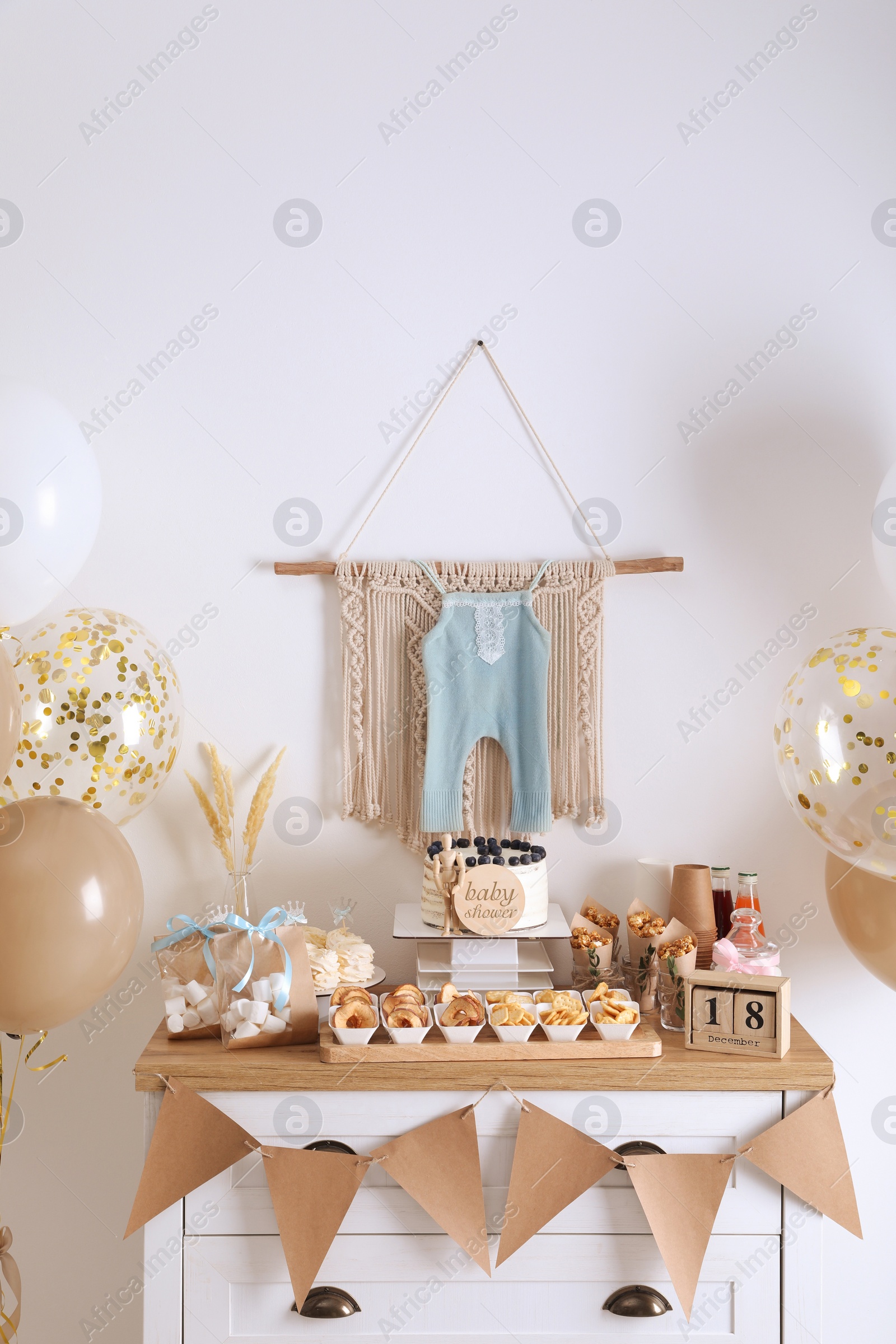 Photo of Baby shower party. Different delicious treats on white wooden chest of drawers and decor near light wall