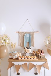 Baby shower party. Different delicious treats on white wooden chest of drawers and decor near light wall