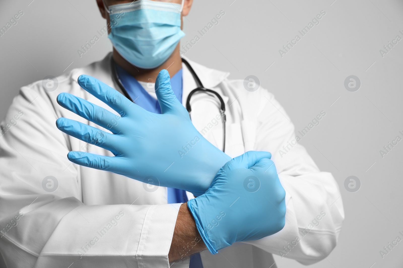 Photo of Doctor in protective mask putting on medical gloves against light grey background, closeup