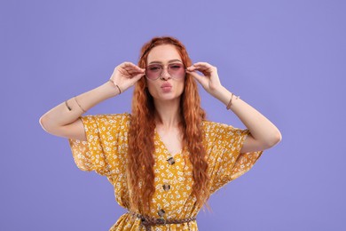Photo of Stylish young hippie woman in sunglasses sending air kiss on violet background