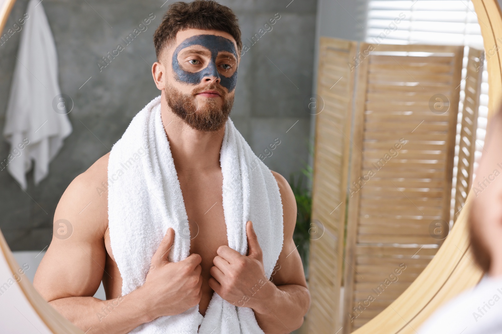 Photo of Handsome man with clay mask on his face near mirror in bathroom