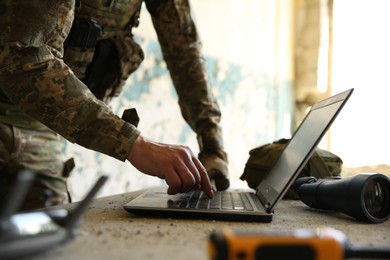 Photo of Military mission. Soldier in uniform using laptop at table inside abandoned building, closeup