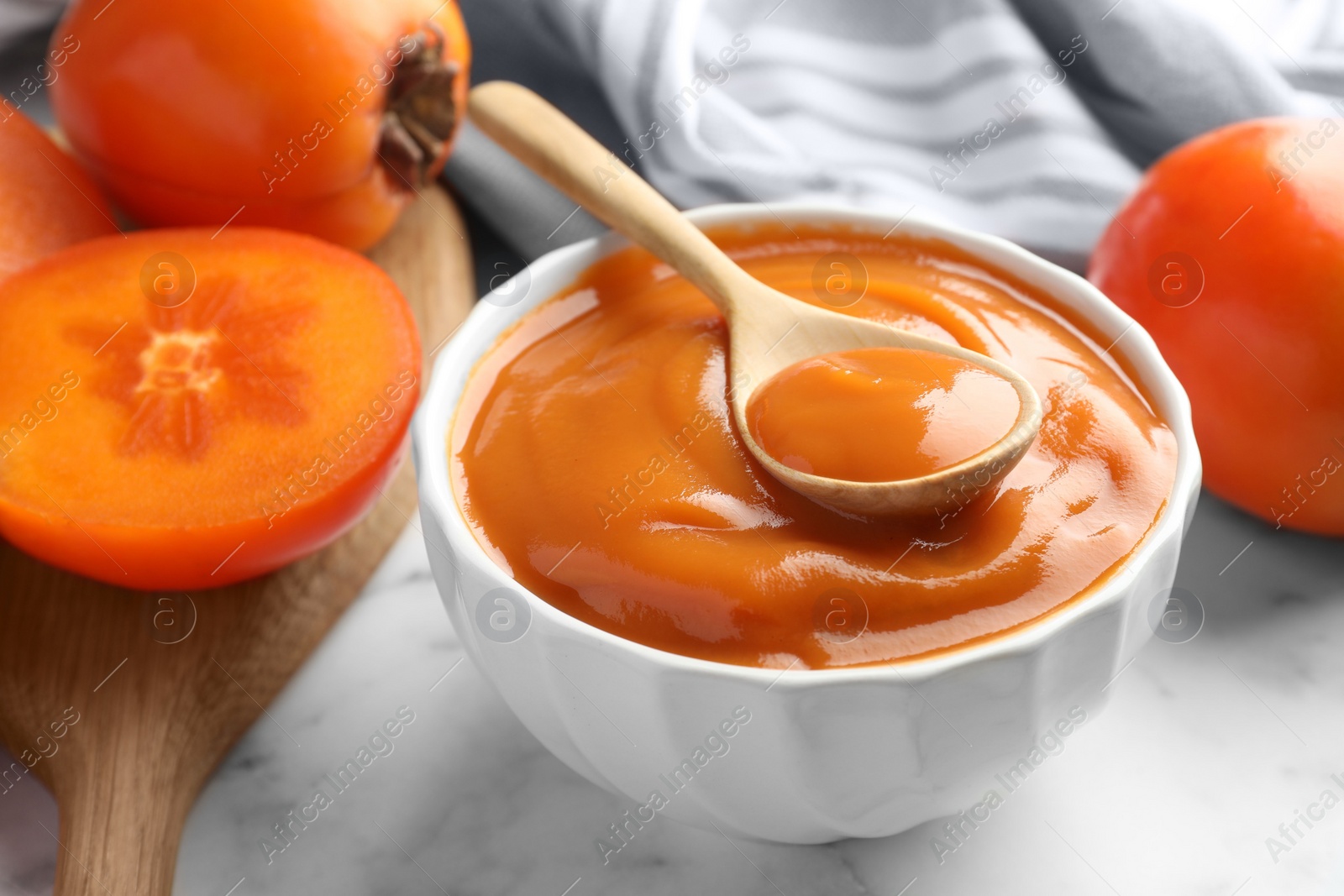 Photo of Delicious persimmon jam and fresh fruits on white marble table, closeup