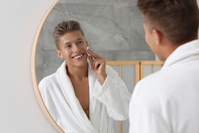 Photo of Handsome young man cleaning face with cotton pad near mirror in bathroom