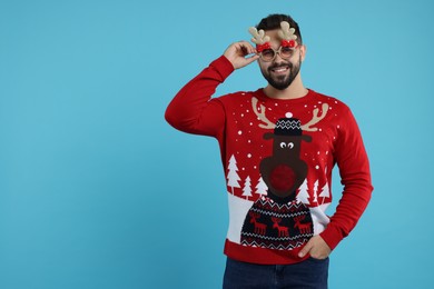 Photo of Happy young man in Christmas sweater and funny glasses on light blue background. Space for text