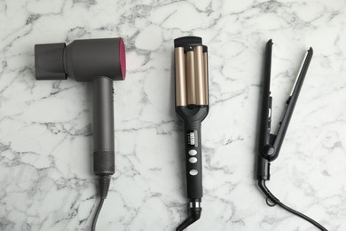 Hair dryer, straightener and triple curling iron on white marble background, flat lay