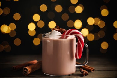 Photo of Glass cup of tasty cocoa with marshmallows and Christmas candy cane on wooden table against blurred festive lights