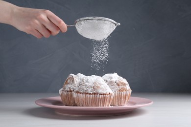 Photo of Woman with sieve sprinkling powdered sugar onto muffins at white wooden table, closeup
