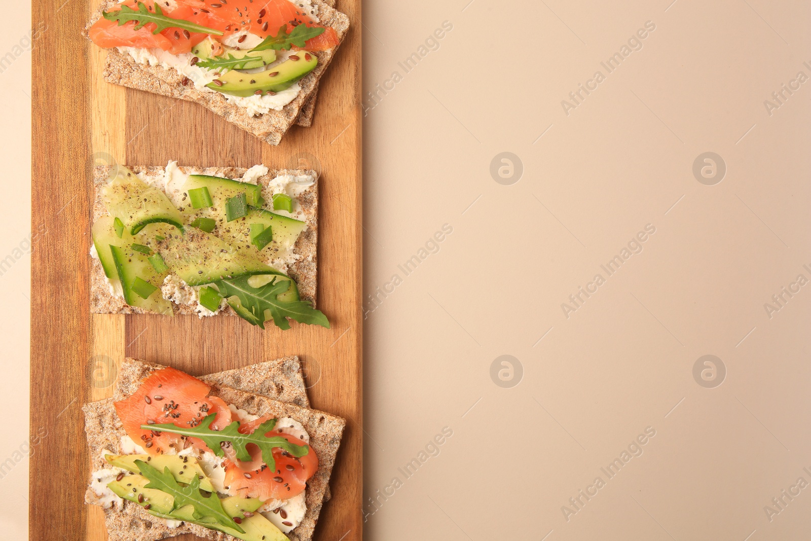 Photo of Fresh crunchy crispbreads with cream cheese, cucumber, green onion, salmon and arugula on beige table, top view. Space for text