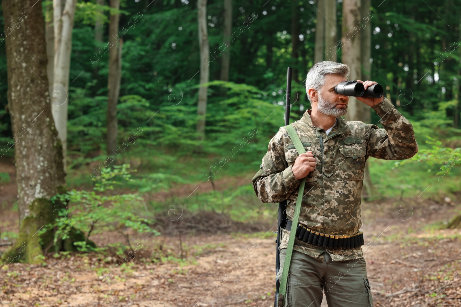 Photo of Man with hunting rifle looking through binoculars in forest. Space for text