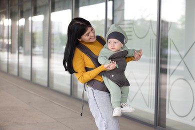 Mother holding her child in sling (baby carrier) near building outdoors